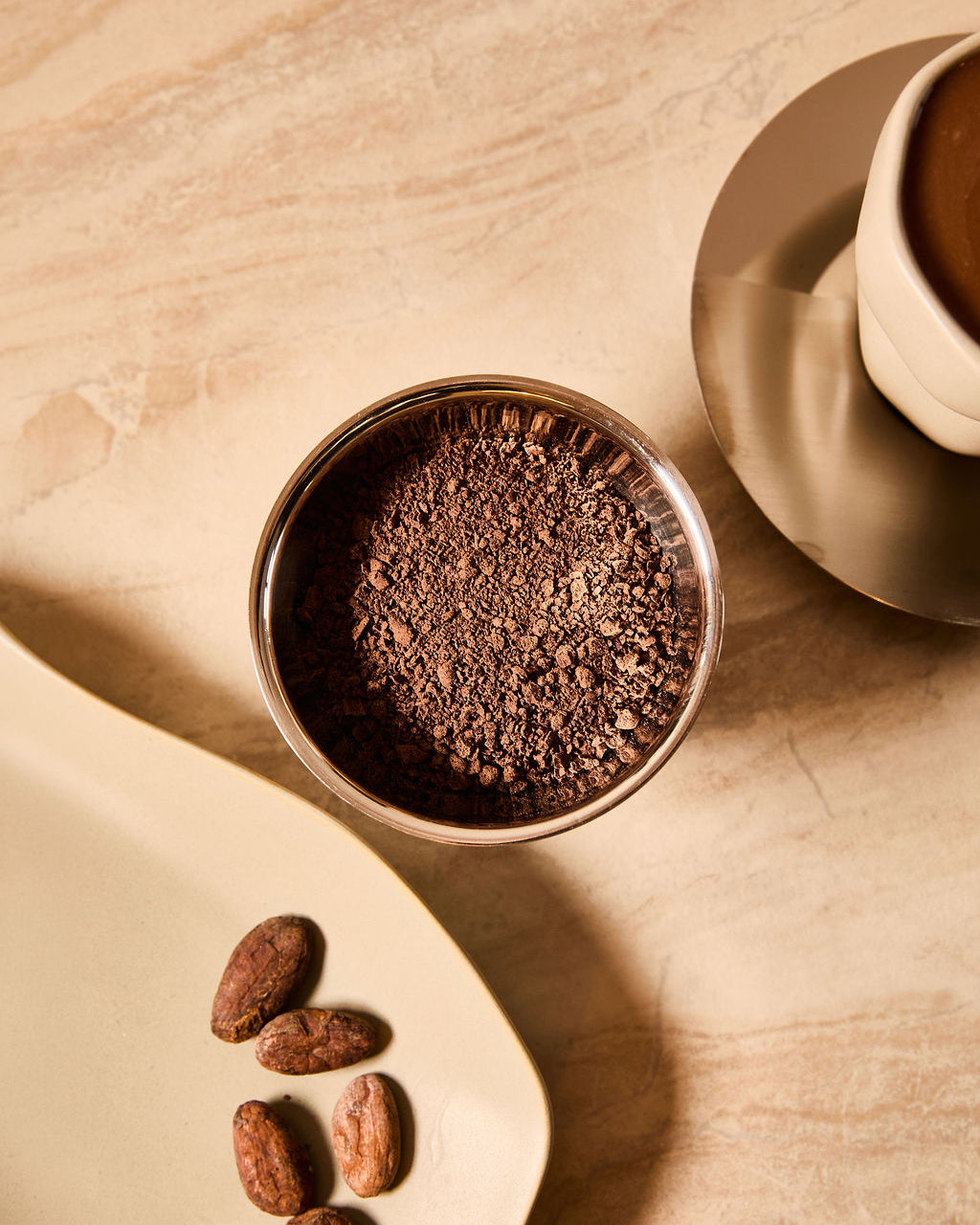 Bowl of cacao powder with raw cacao beans on a tray next to a cup of ceremonial cacao.