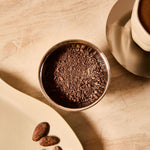 Bowl of cacao powder with raw cacao beans on a tray next to a cup of ceremonial cacao.