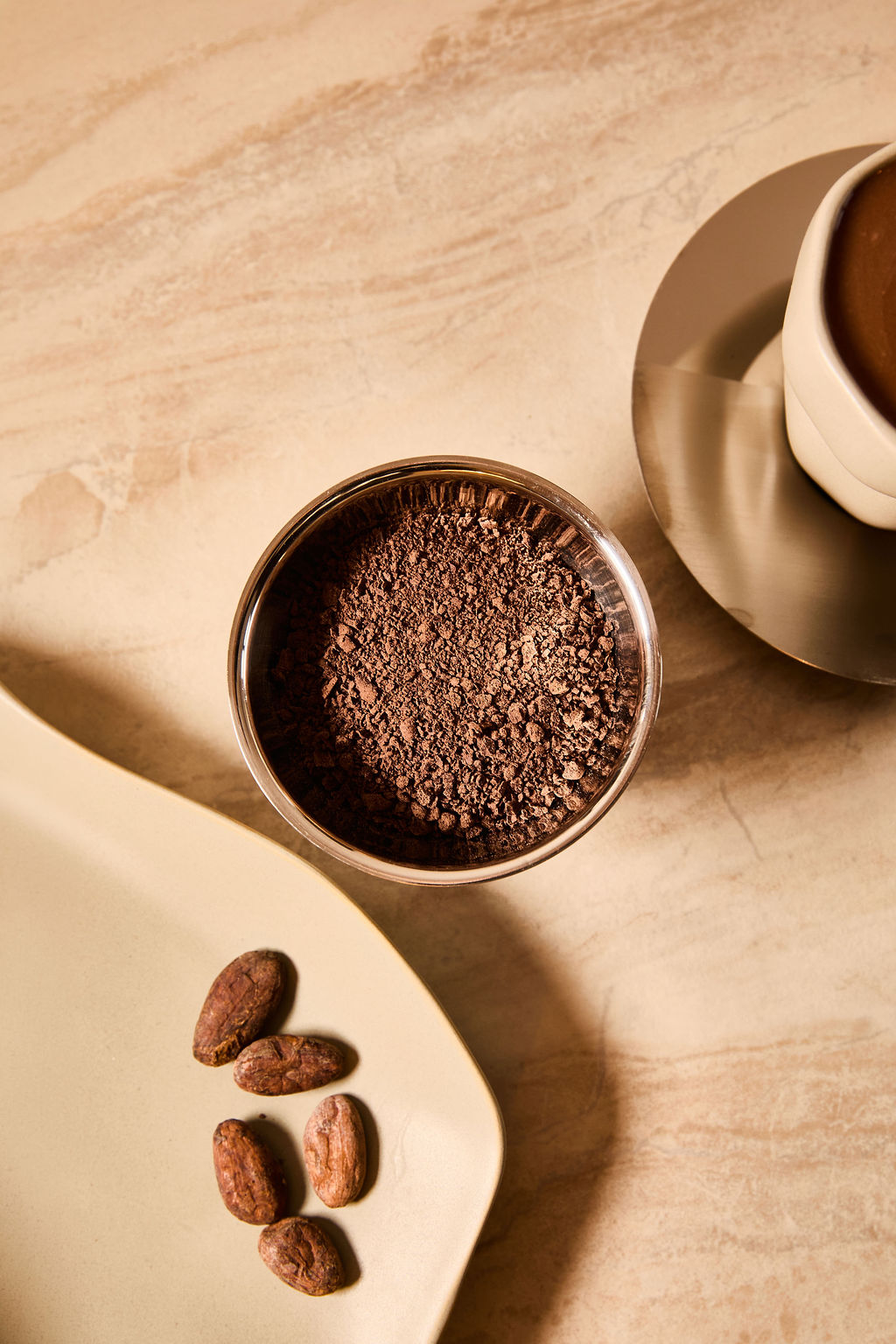 Bowl of cacao powder with raw cacao beans on a tray next to a cup of ceremonial cacao.