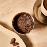 Bowl of cacao powder with raw cacao beans on a tray next to a cup of ceremonial cacao.