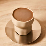 Minimalist beige ceramic cup filled with ceremonial cacao placed on a round brass coaster.