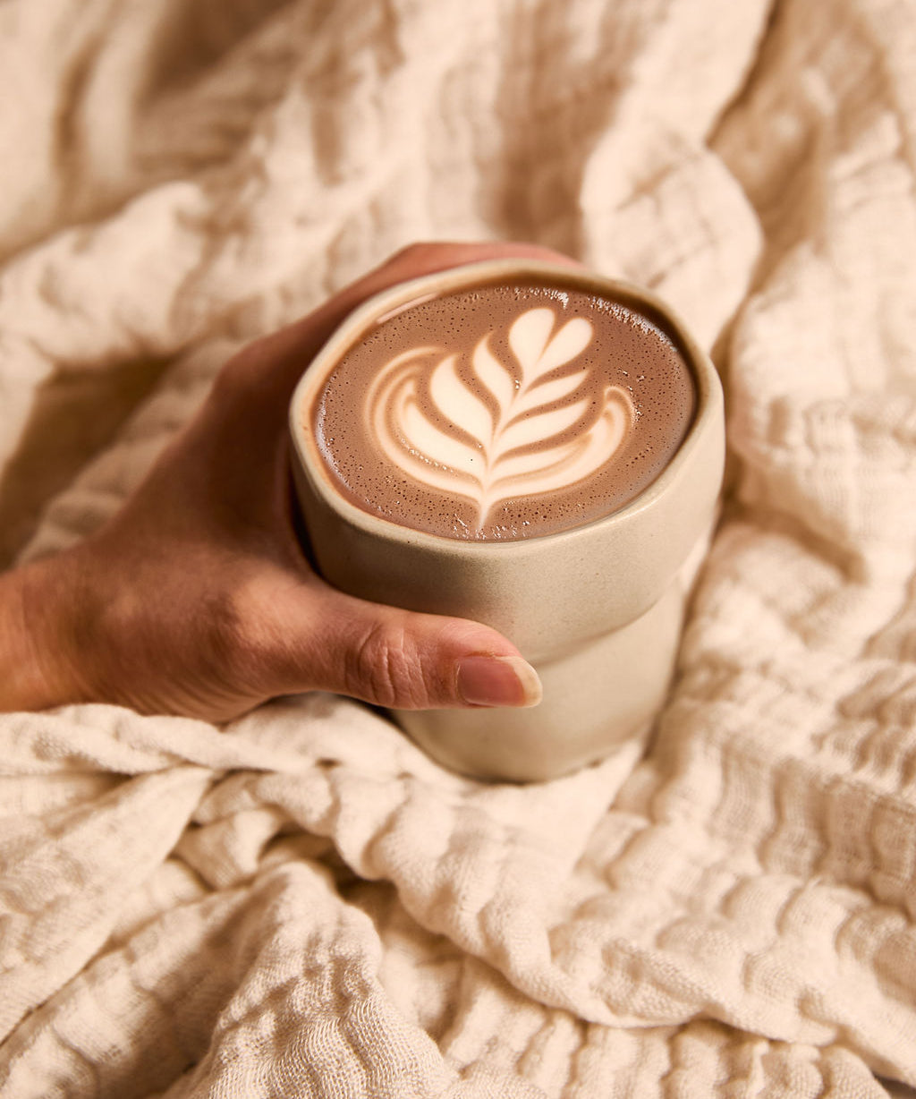 Hand holding a warm cup of ceremonial cacao with latte art, wrapped in a soft beige blanket.