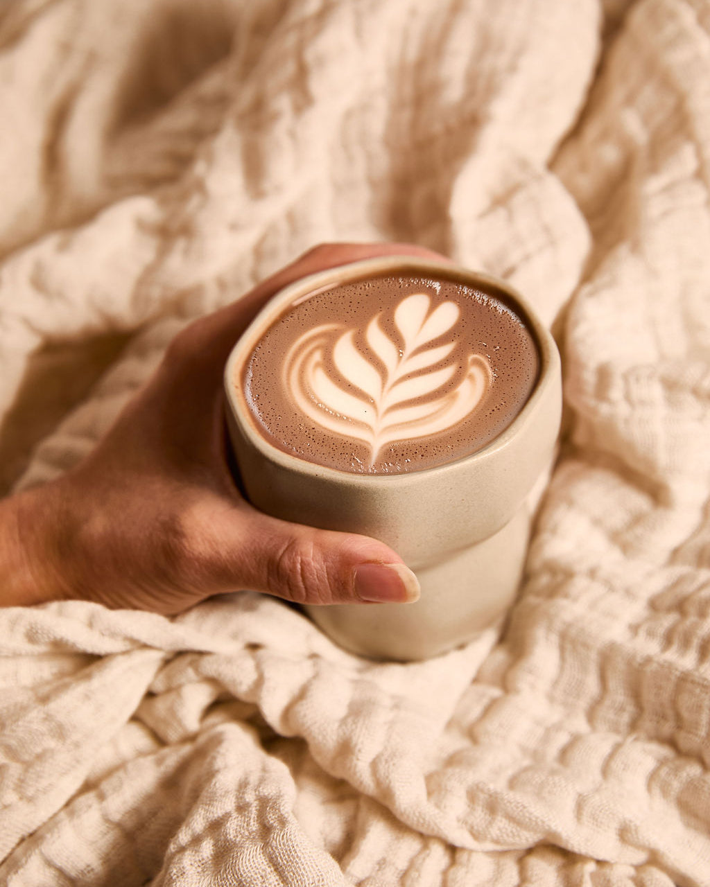 Hand holding a warm cup of ceremonial cacao with latte art, wrapped in a soft beige blanket.