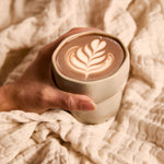 Hand holding a warm cup of ceremonial cacao with latte art, wrapped in a soft beige blanket.