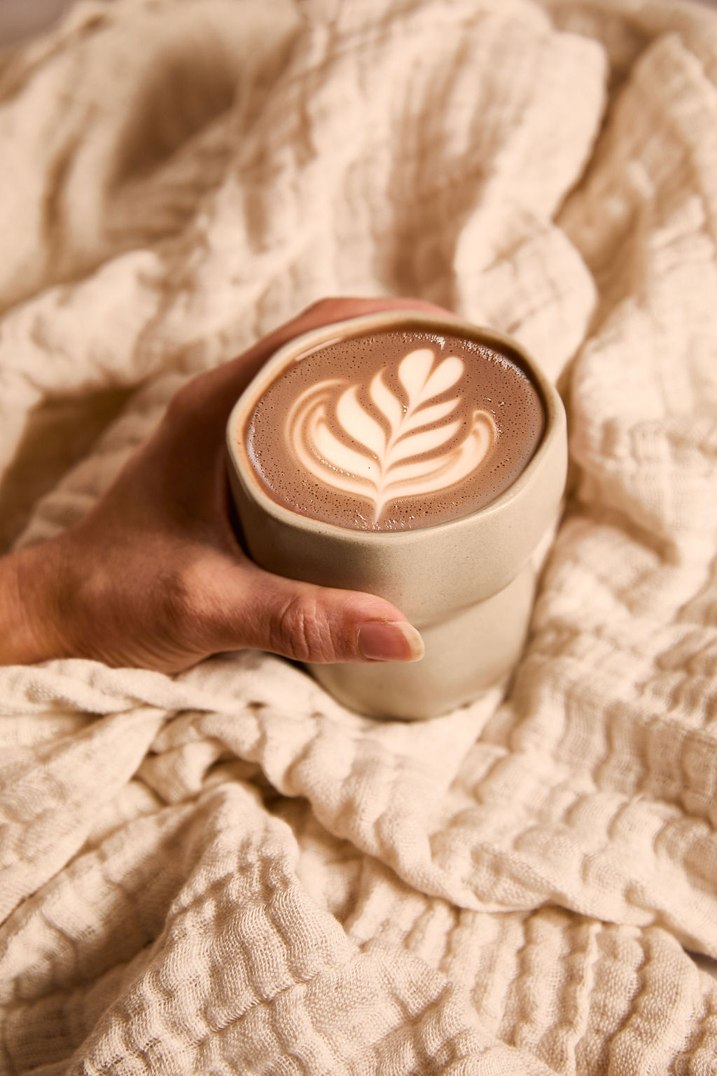 Hand holding a warm cup of ceremonial cacao with latte art, wrapped in a soft beige blanket.
