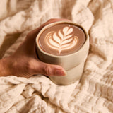 Hand holding a warm cup of ceremonial cacao with latte art, wrapped in a soft beige blanket.