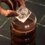 Hand placing a large ice cube into a glass of iced ceremonial cacao on a dark marble surface.