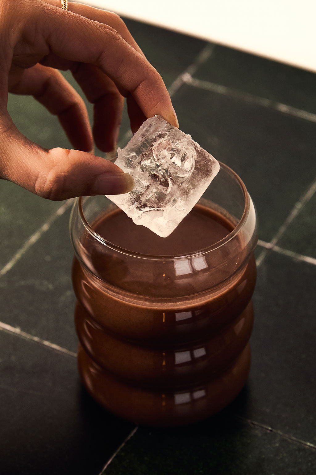 Hand placing a large ice cube into a glass of iced ceremonial cacao on a dark marble surface.