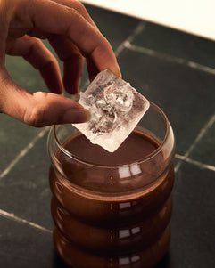 Hand placing a large ice cube into a glass of iced ceremonial cacao on a dark marble surface.